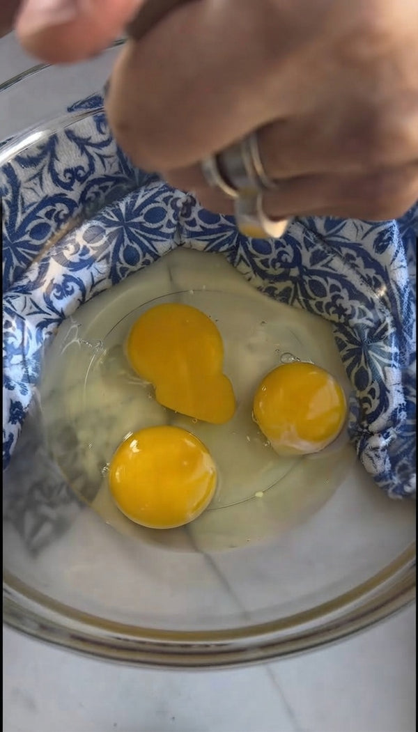 Crack fresh eggs into a mixing bowl lined with a patterned kitchen towel for stability, ensuring the yolks remain intact alongside the egg whites.